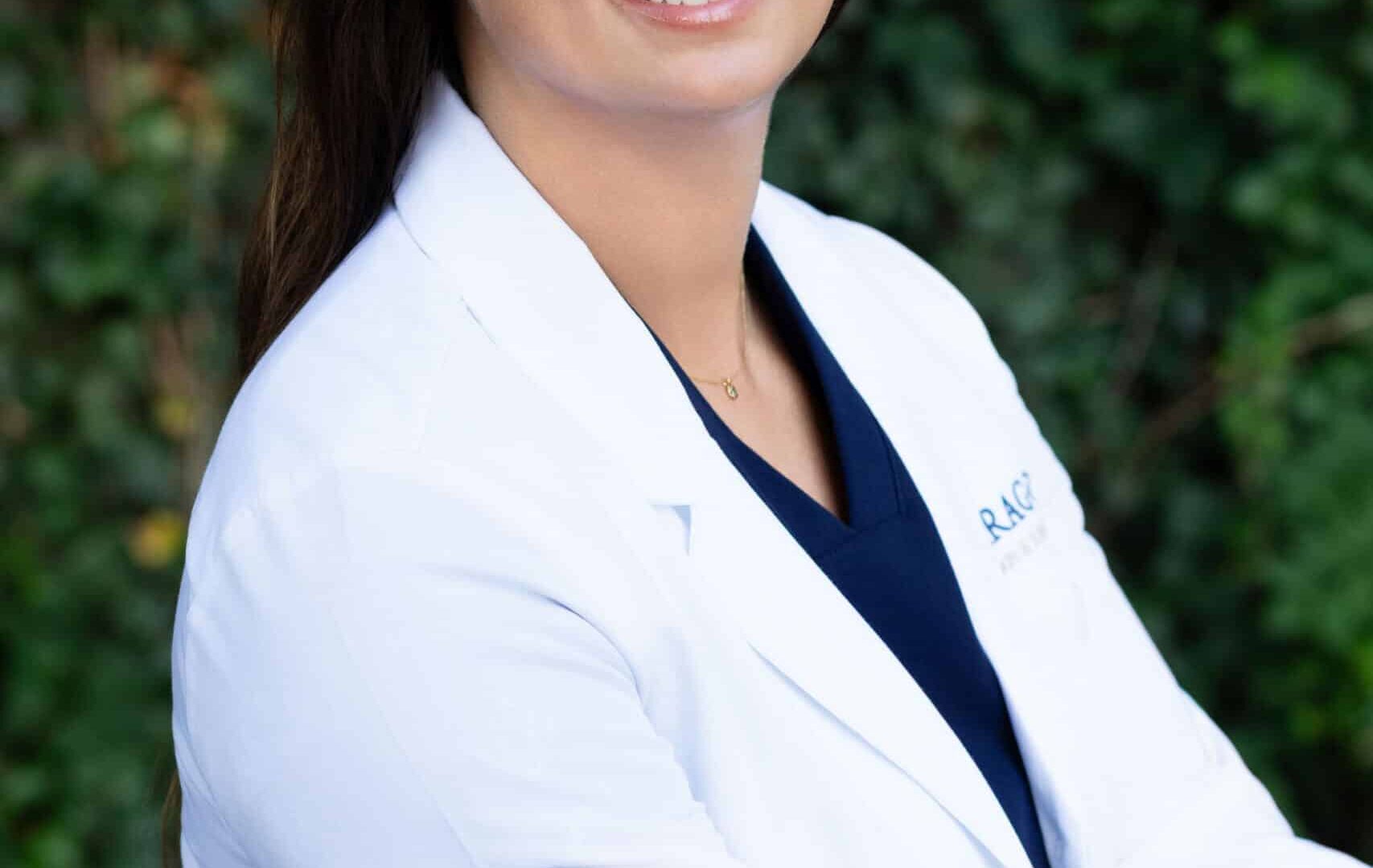 Dermatologist in white coat smiling outdoors with greenery background